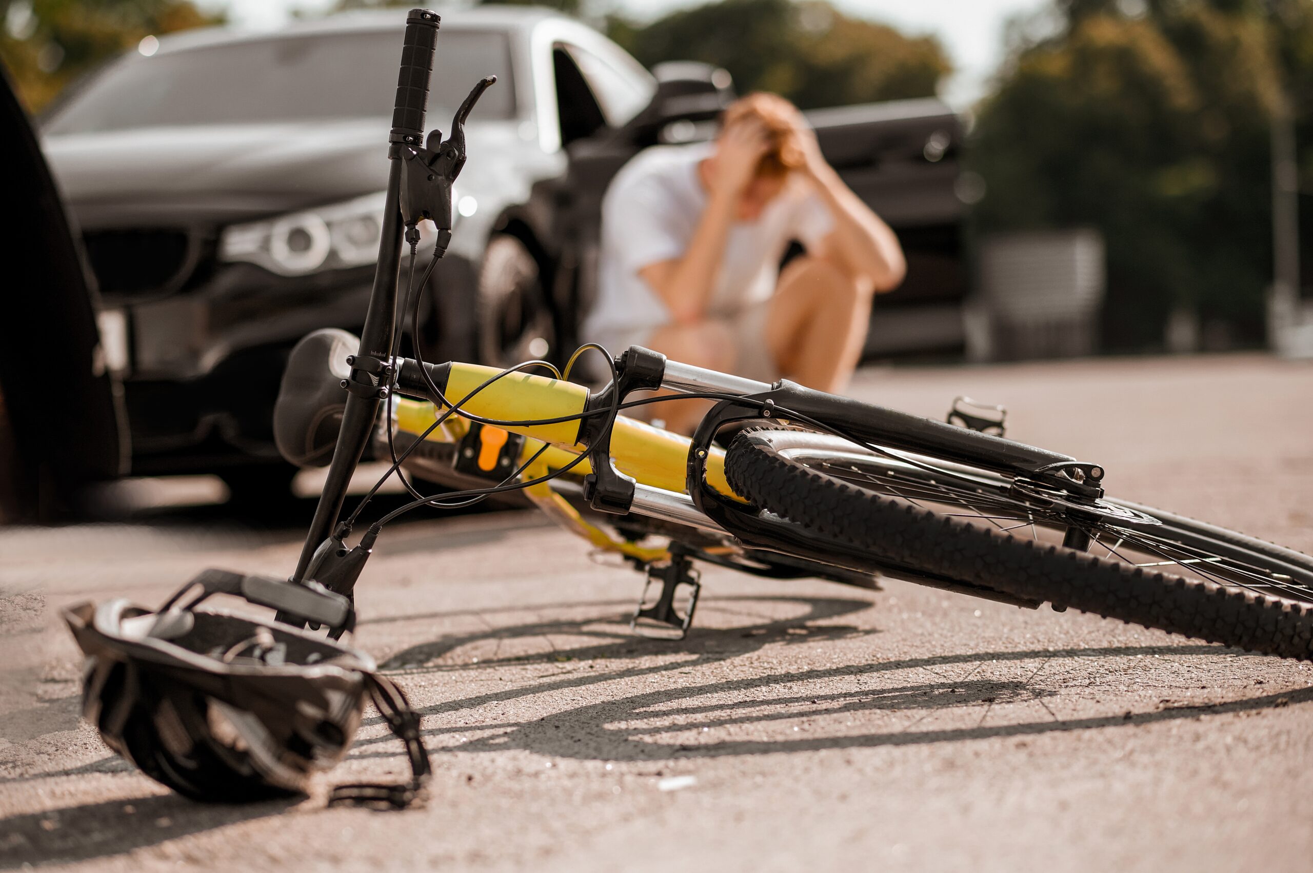 picture of yellow bike and helmet turned on its side in the middle of a street and in the background a black car with its driver kneeling down beside the car with his head in his hands