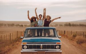 Three women happily riding in the bed of a truck on a rural dirt road.