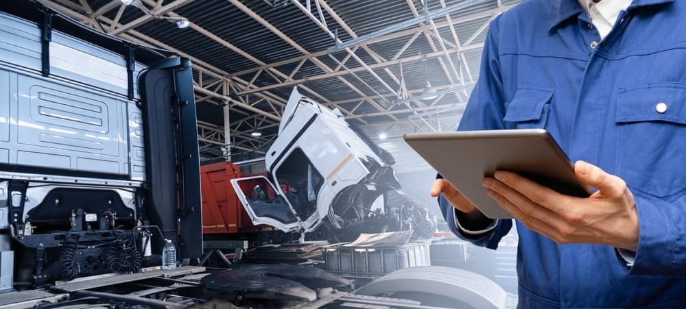 A man in blue overalls stands with a tablet, inspecting a truck for maintenance.