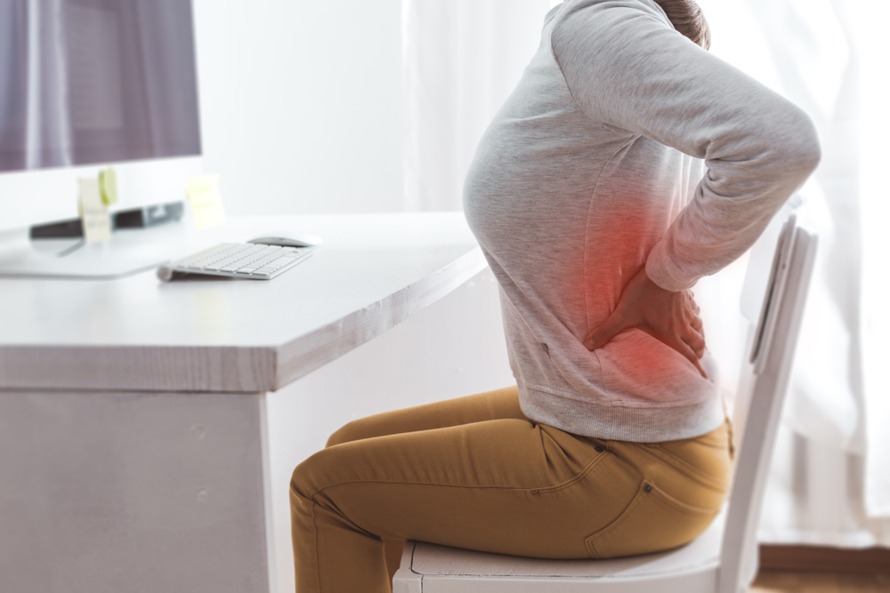 woman holding her lower back in pain sits at a white chair behind white desk