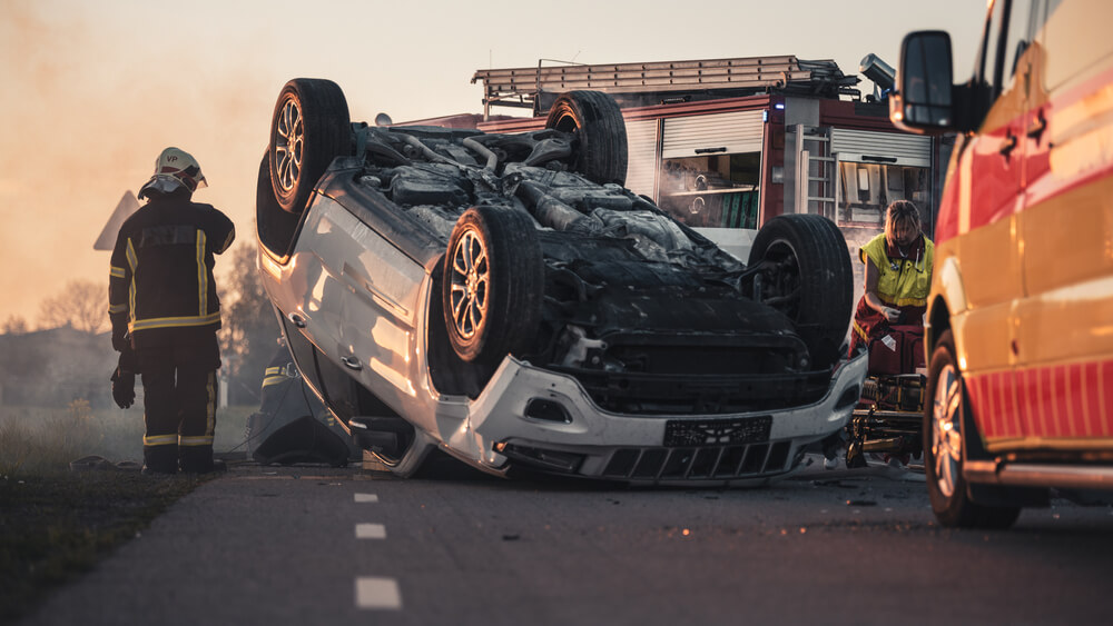 Traffic Accident Rollover Smoking and Burning Vehicle Lying on its Roof in the Middle of the Road after Collision.